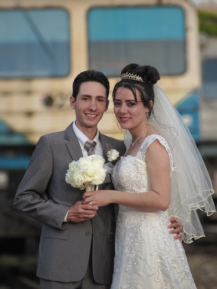 A happy couple in wedding attire poses outdoors against a blurred background.